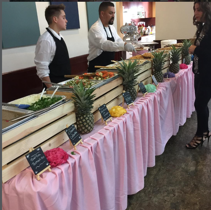 Línea de buffet con mantelería rosa, cajas de madera, piñas y pizarrones con los nombres de las salsas