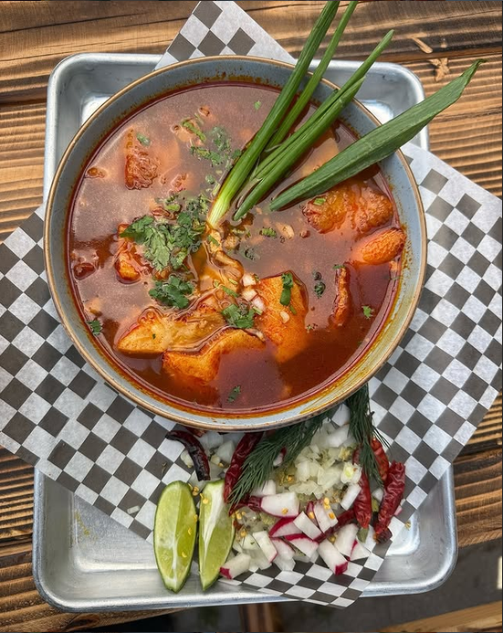 Tazón de menudo en caldo rojo con cebolla, cilantro, rábano y limón