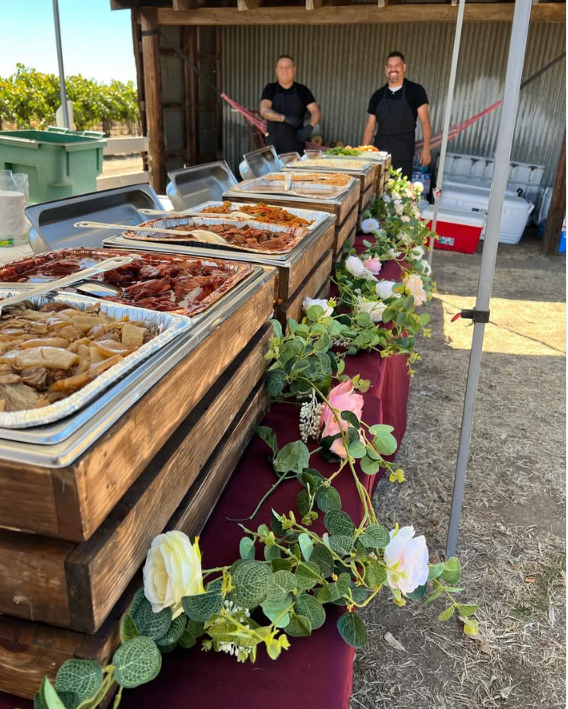Línea de buffet al aire libre sobre cajas de madera, decorada con guirnalda de rosas, en un evento en el campo