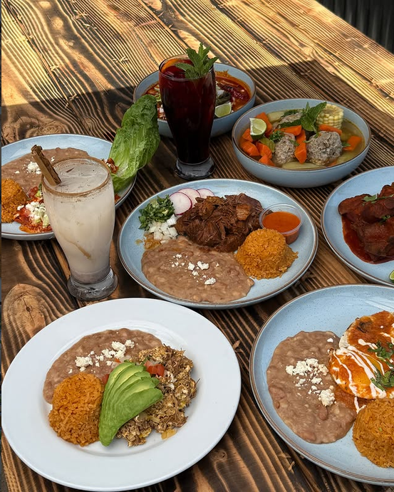 Mesa de madera con varios platillos mexicanos, arroz, frijoles, sopa de albóndigas y aguas frescas